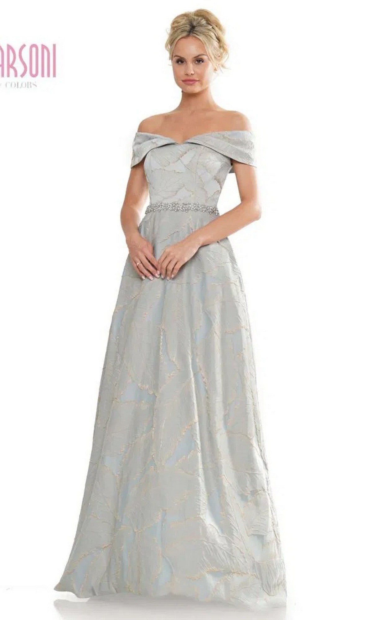 A woman stands with hands clasped in front, wearing the Marsoni MV1265 Long Formal Dress—an elegant light grey off-the-shoulder gown with a fitted bodice, subtle pattern, and beaded waistline.